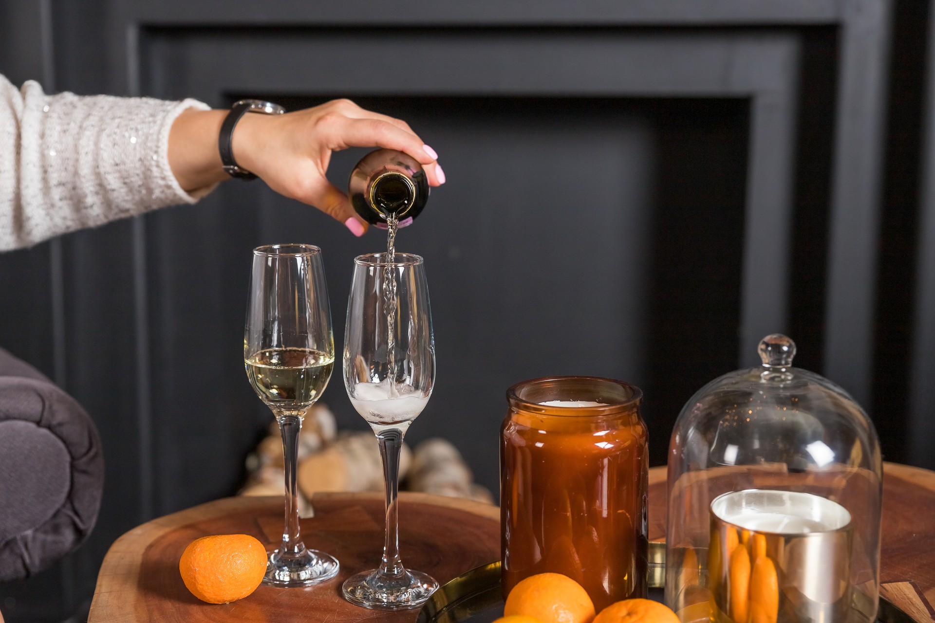 The woman's hand pours champagne from a bottle into glass, tangerines on a table against blurred black background. New year celebration party.Winter holidays celebration